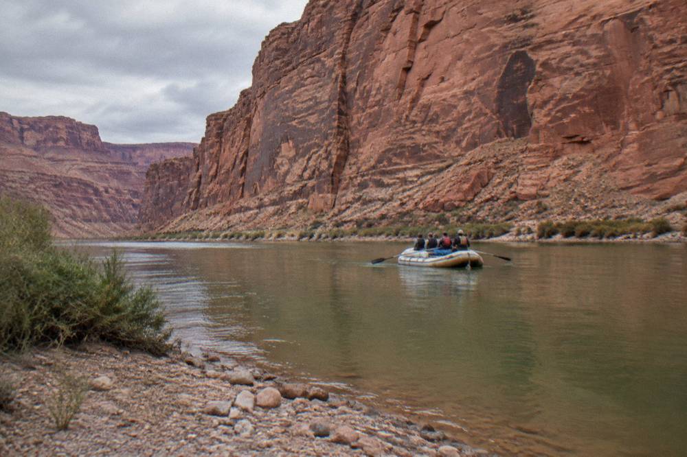 Motorized raft drifting on flat water toward a tall red rock canyon wall