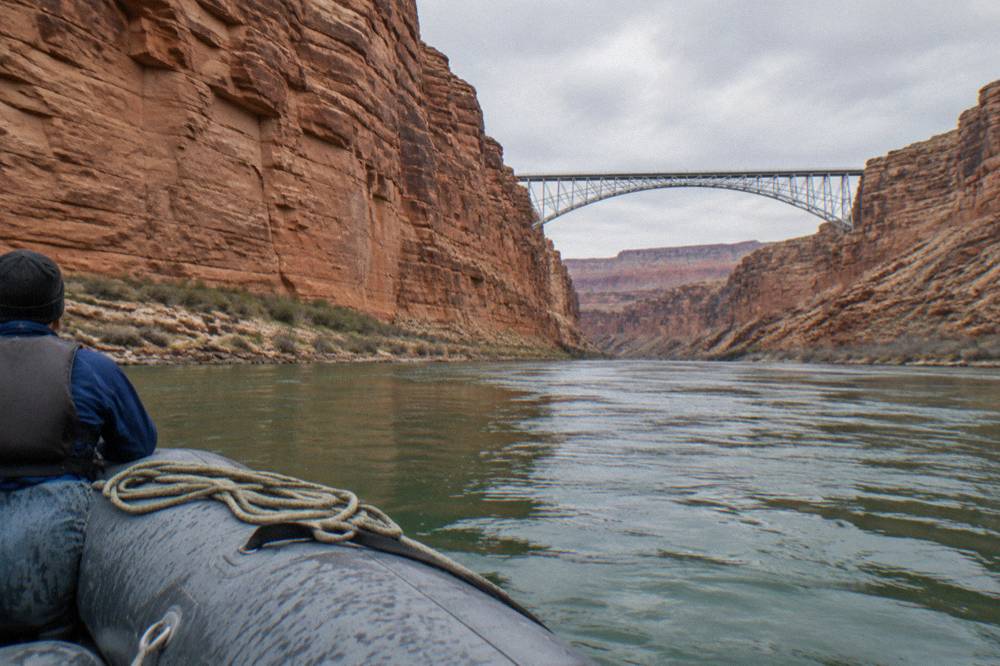 A motor raft on green flat water passing beneath high orange sandstone cliffs
