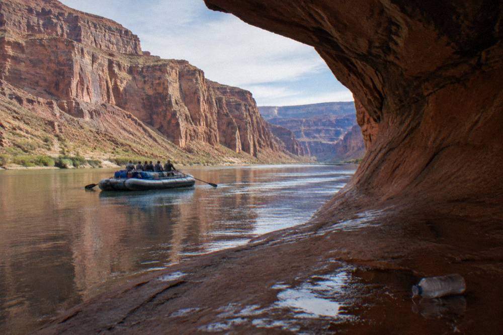 Passengers on a flat-bottomed raft floating past a curved sandstone cliff above still water