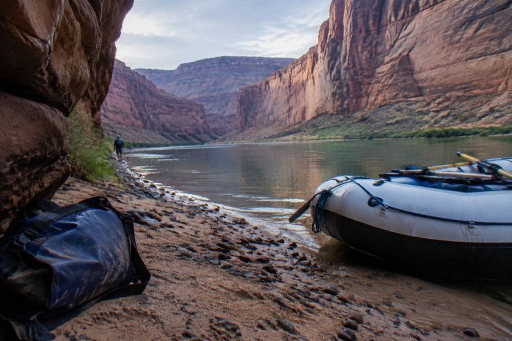 Raft resting on a sandy beach beside green river water at the base of tall cliffs
