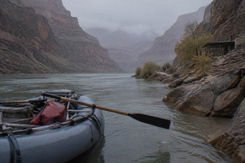 Rowed oar raft mid-river with a canyon wall towering above in morning light