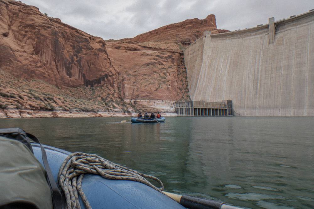 A guided raft passing near the base of a massive concrete dam wall with canyon cliffs rising above