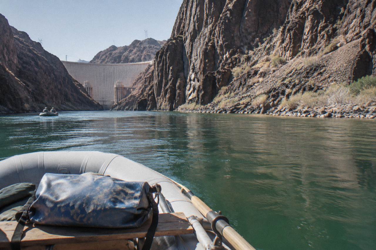 A raft floating on calm green water beneath tall volcanic cliffs in Black Canyon below Hoover Dam
