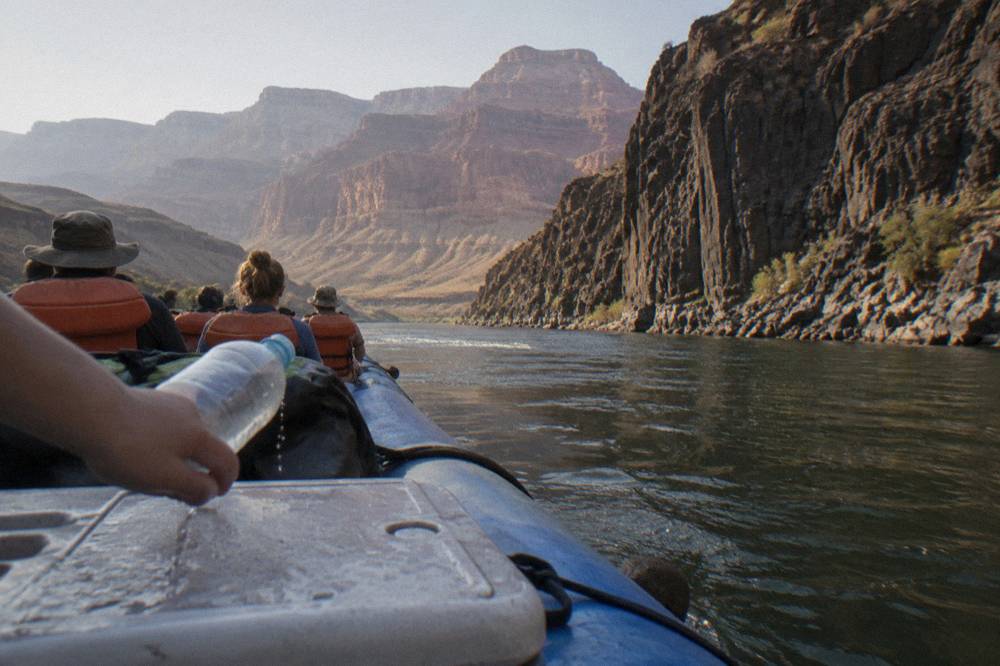 Raft passengers in life jackets floating past tall dark canyon cliffs