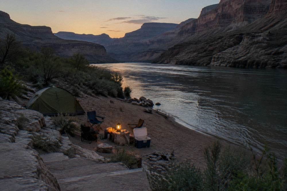 Camping gear arranged on a sandy river beach beside calm water at dusk