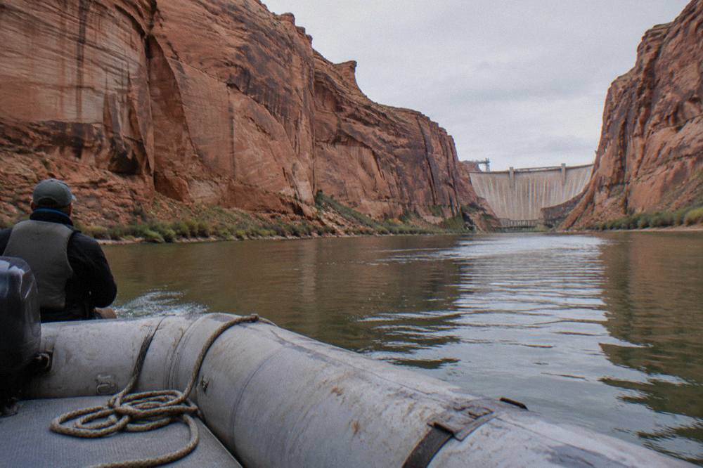 A motorized raft drifting on calm green water between tall red sandstone canyon walls.