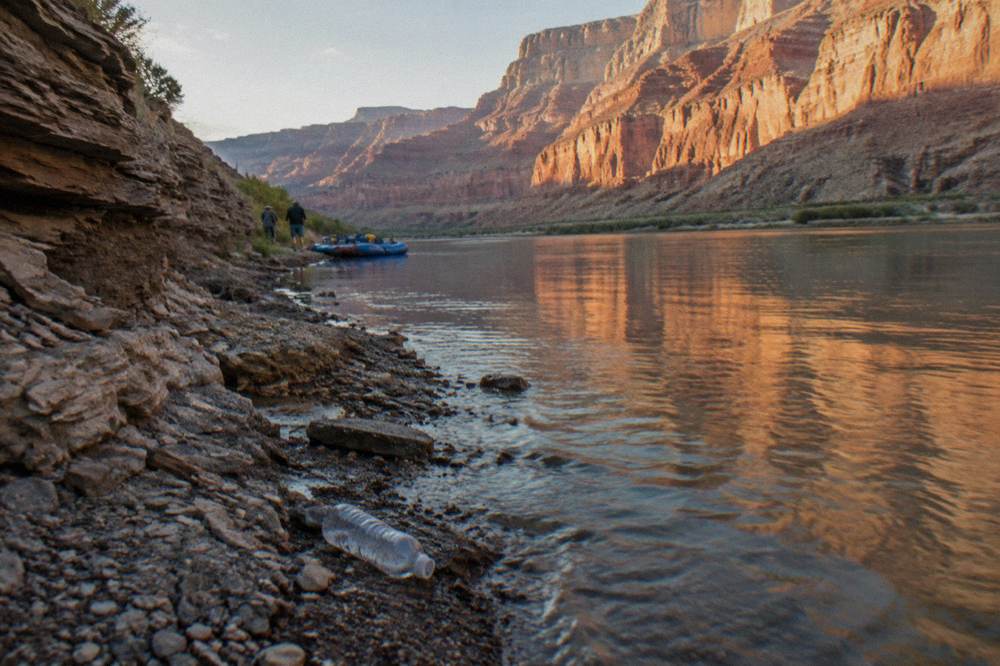 A raft passengers view of low sandstone walls glowing warm in late afternoon light above calm water.
