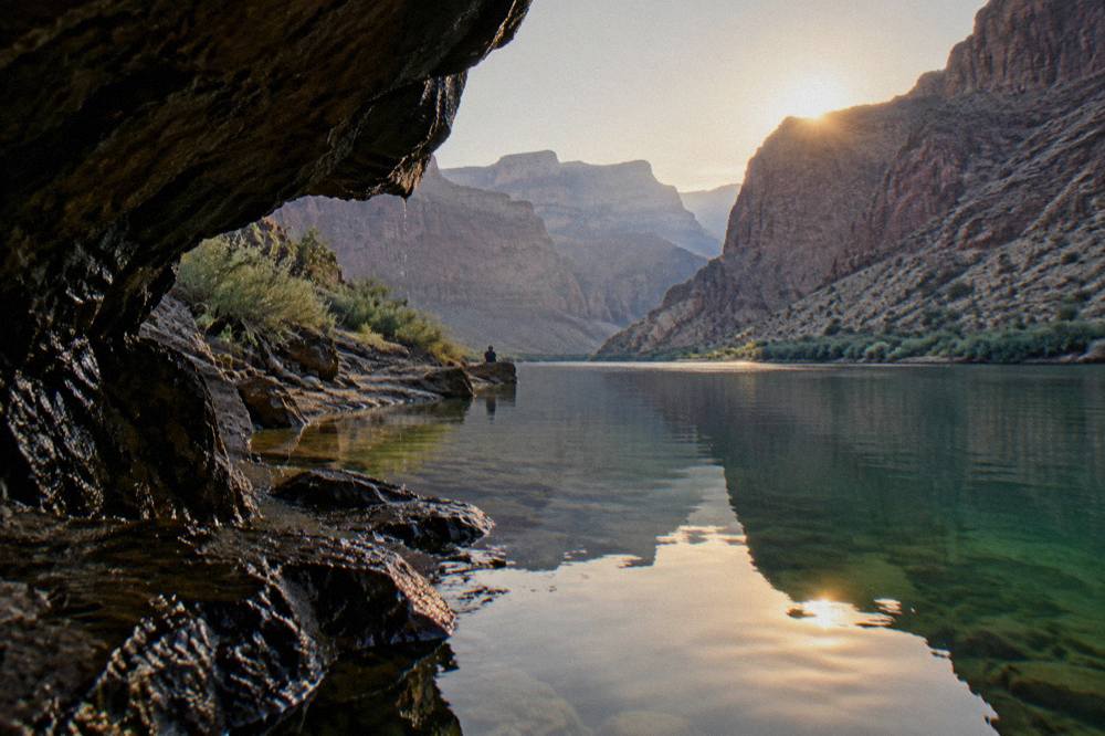 A low river cave opening with green water reflecting light at the base of dark canyon walls
