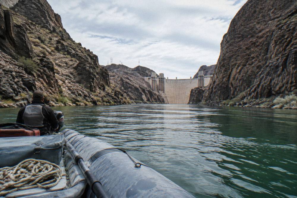 A guided motorized raft drifting along a flat green river beneath steep canyon cliffs