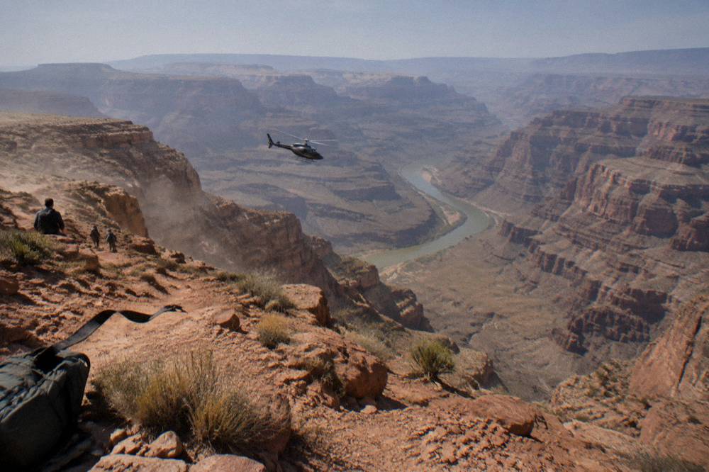 A helicopter flying low over a wide desert canyon toward a distant river bend