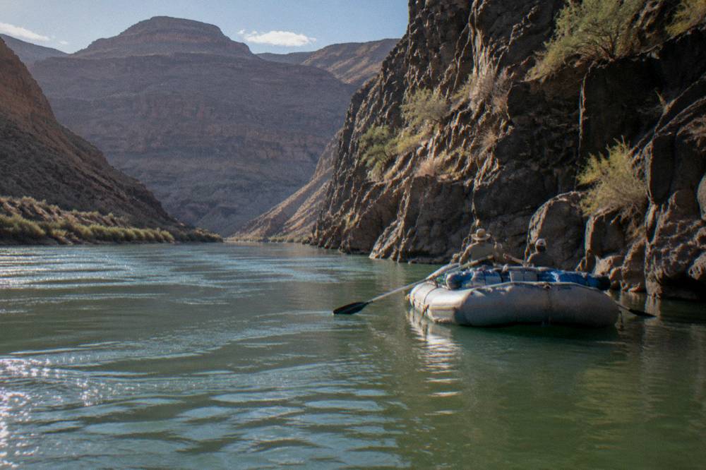 Raft floating on green river water beside tall rock cliffs with sparse desert shrubs