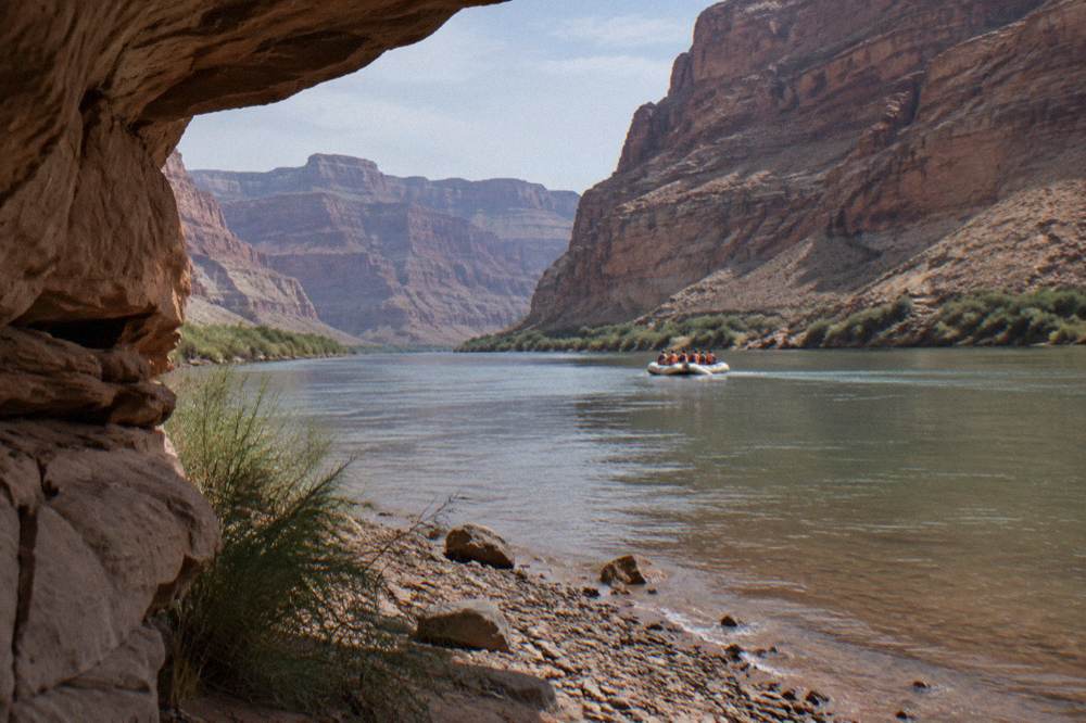 A motorized raft carrying passengers along a wide calm river in a deep canyon