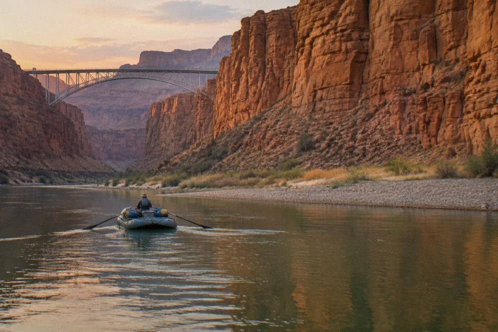 A guide rowing an oar raft through a calm green stretch of river beneath canyon cliffs