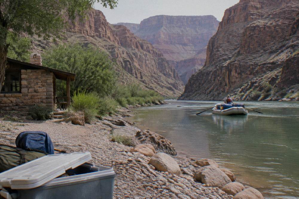 A guide in a life jacket steering an oar raft through a calm stretch of canyon river