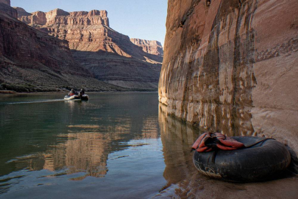 A motorized raft drifting on glassy green water beside a sheer sandstone cliff