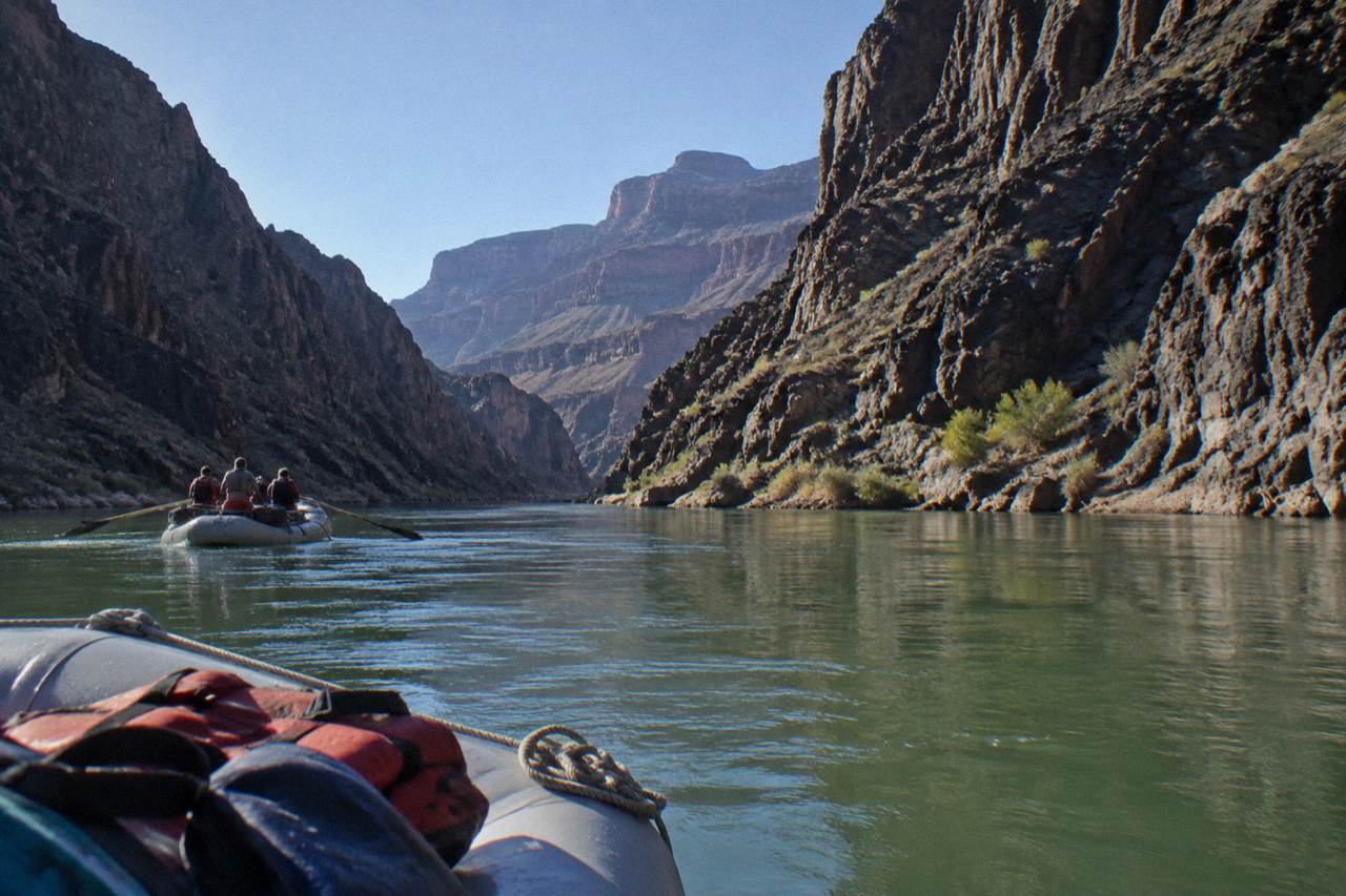 A motorized raft drifting on calm green water beneath tall canyon walls on the Colorado River