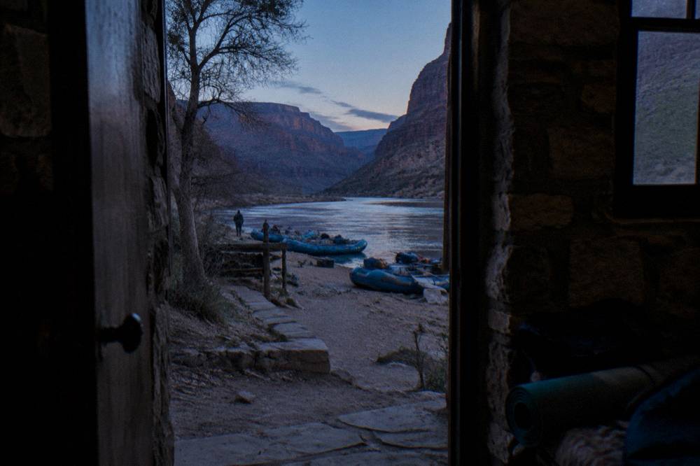 Camping gear arranged on a riverbank beside moored rafts at dusk
