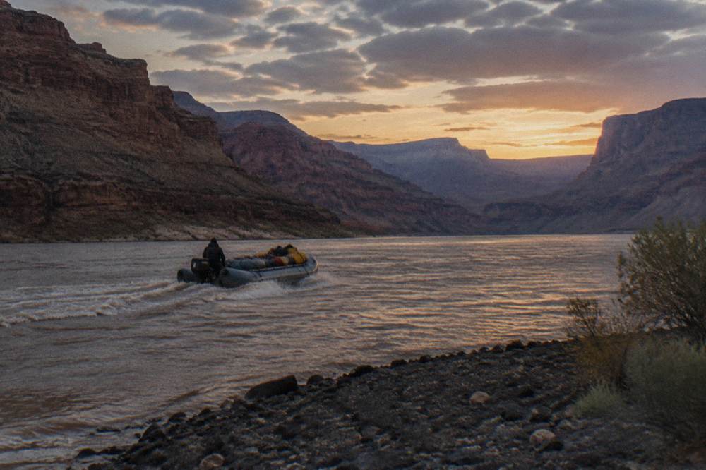 A river guide steering a motorized raft with passengers seated and wearing life jackets