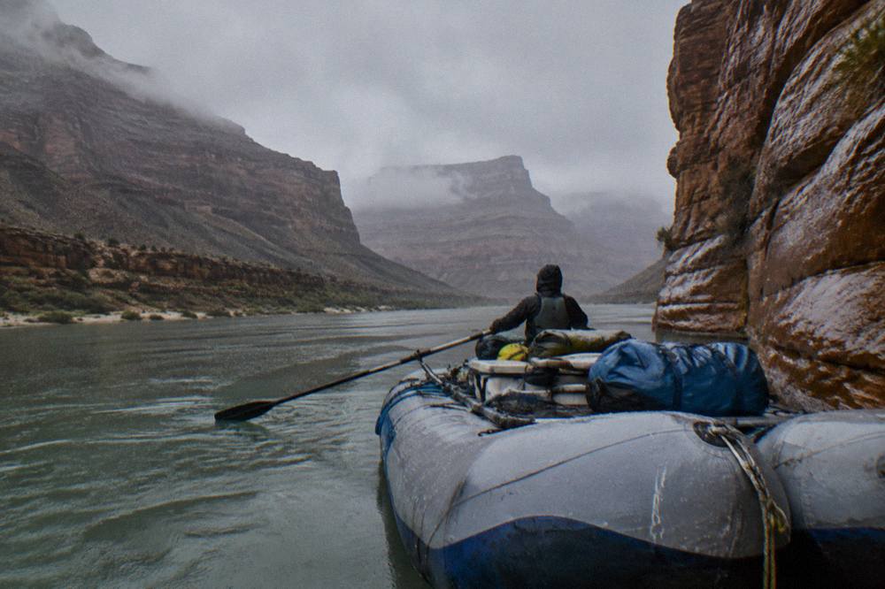 A wide raft with a guide rowing oars on a flat green river between sandstone walls