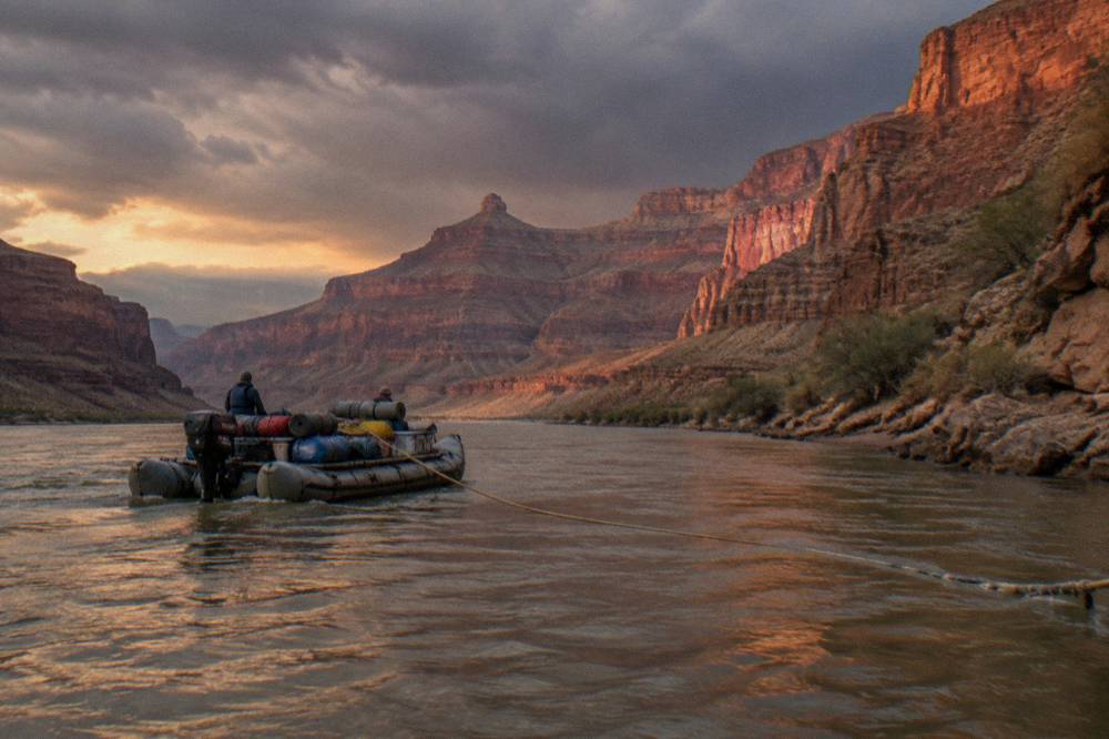A long motorized raft loaded with camping gear heading down a wide river bend at sunset