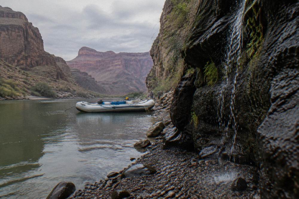 A raft floating near a tall canyon wall with a small water cascade at its base