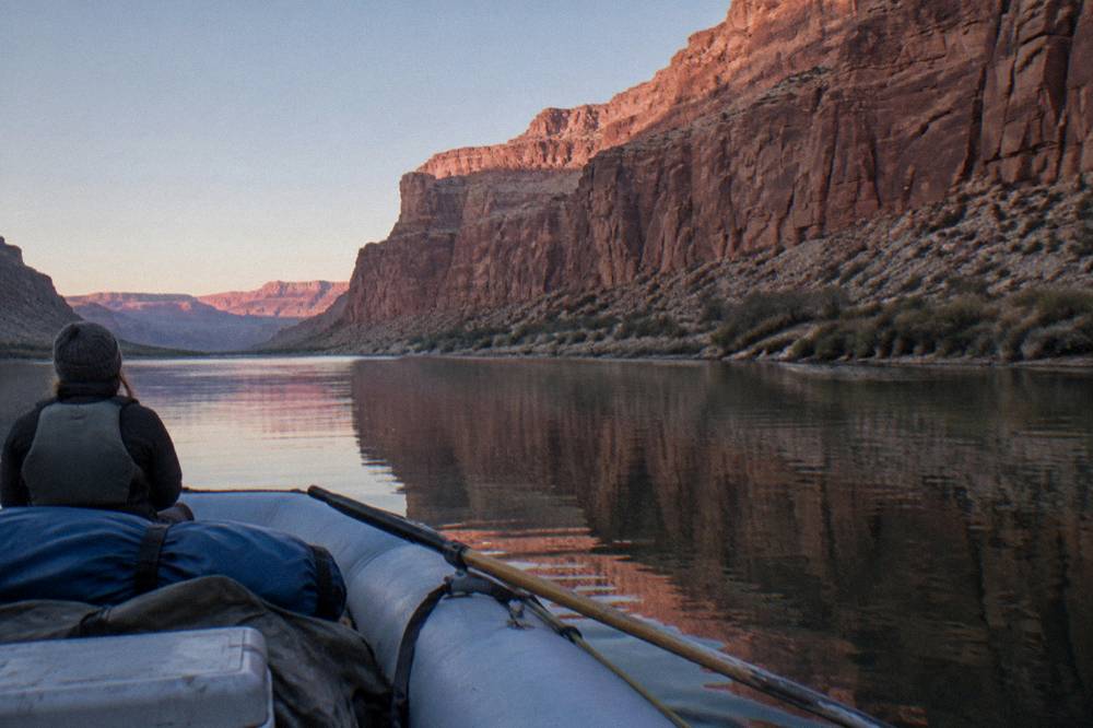 A motor raft drifting on flat water toward a tall sandstone cliff reflected in the river
