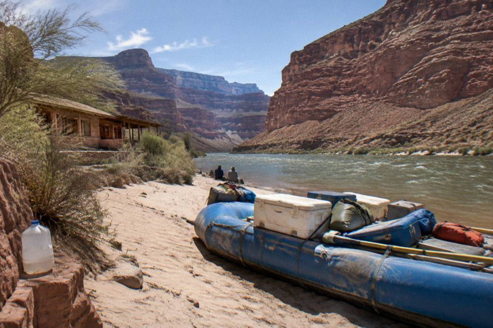 Camping gear and coolers arranged on a large raft pulled onto a sandy beach at the base of a red rock cliff