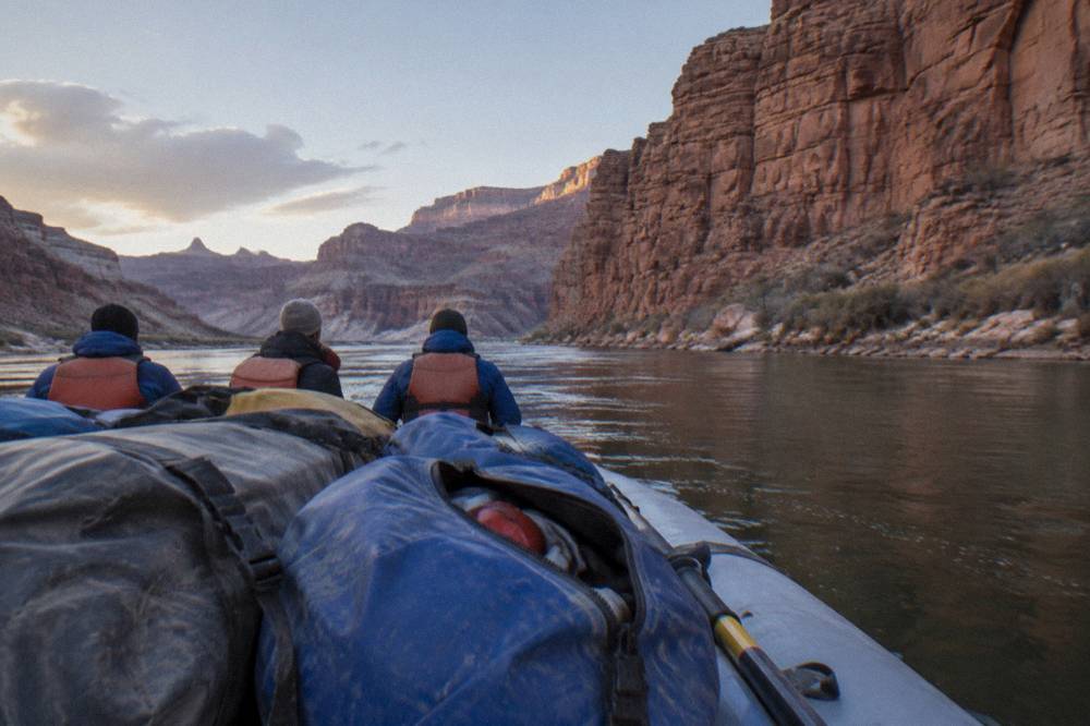Family in life jackets sitting on gear bags aboard a raft in a calm river stretch