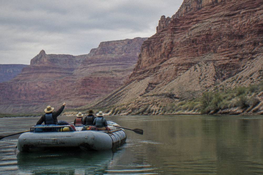 A family sitting in a raft on flat water while a guide points at canyon walls