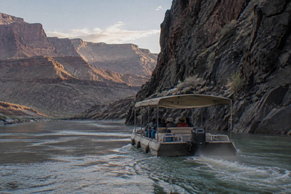 A motorized raft with shaded passengers gliding along a flat green river reach below tall rock walls