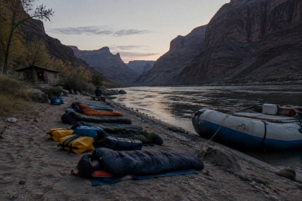 A row of sleeping bags and dry bags arranged on a sandy river beach beside a tied-up raft